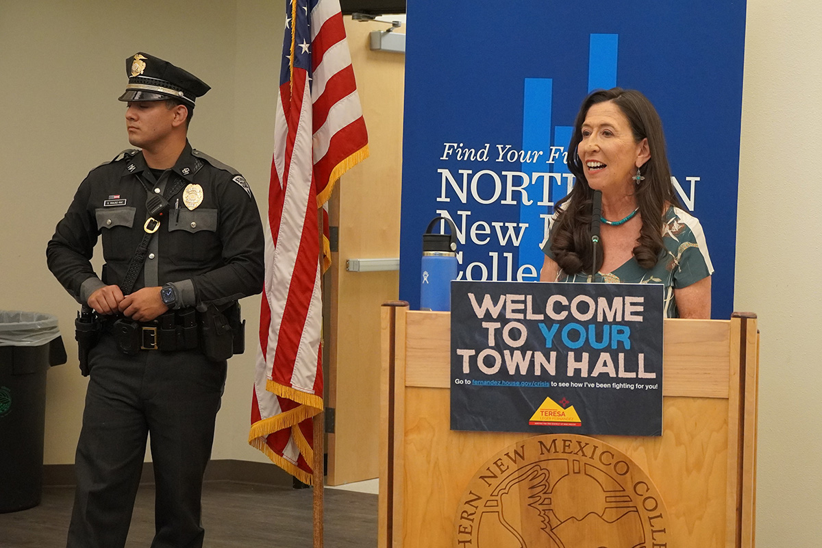 Congresswoman Teresa Leger Fernández at a podium with a sign reading "Welcome to Your Town Hall" with a Northern New Mexico College banner behind her and a State Police officer to her right.